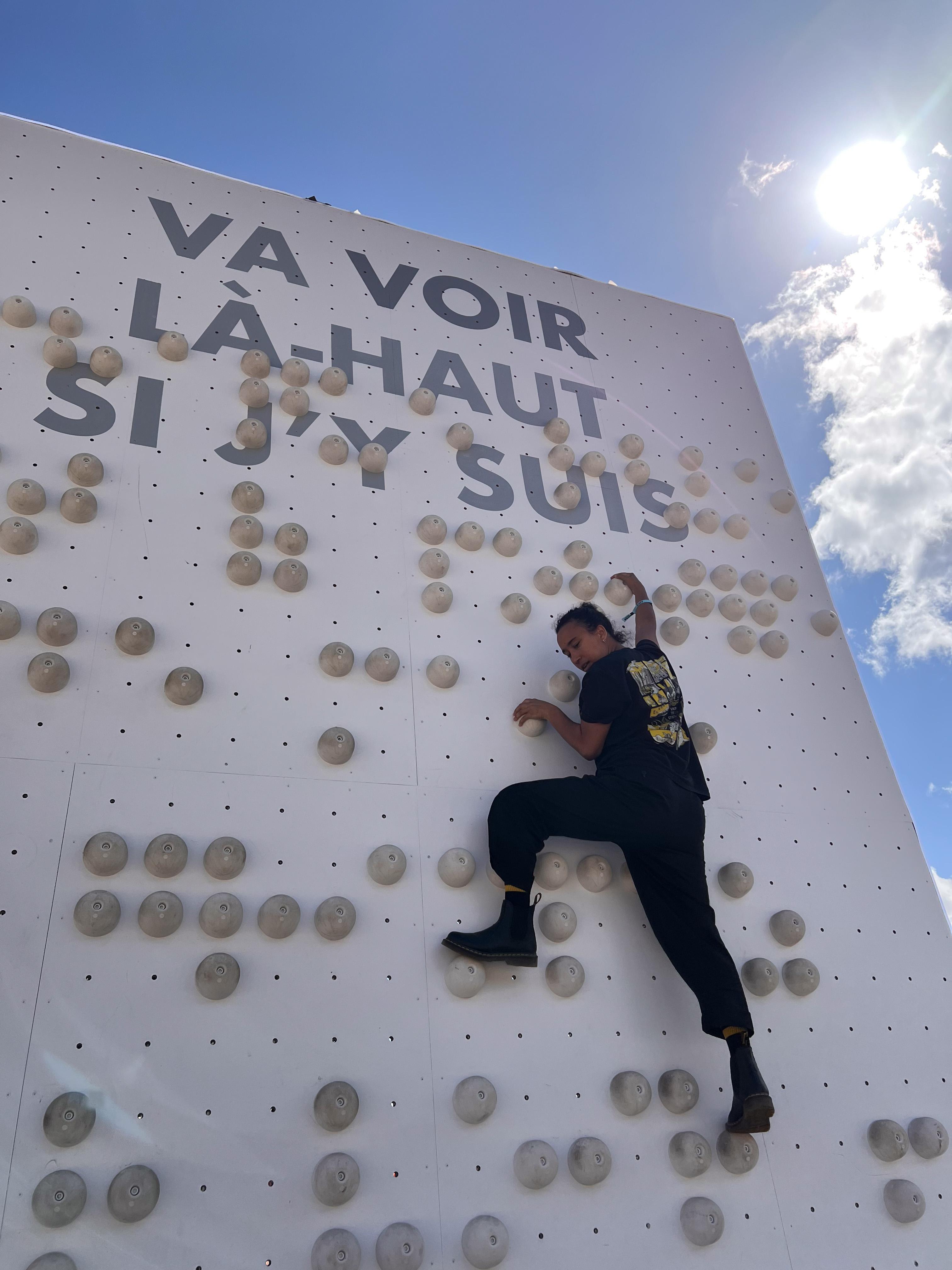 Une personne monte un mur braille, en haut du mur est écrit la phrase "Va voir là-haut si j'y suis"