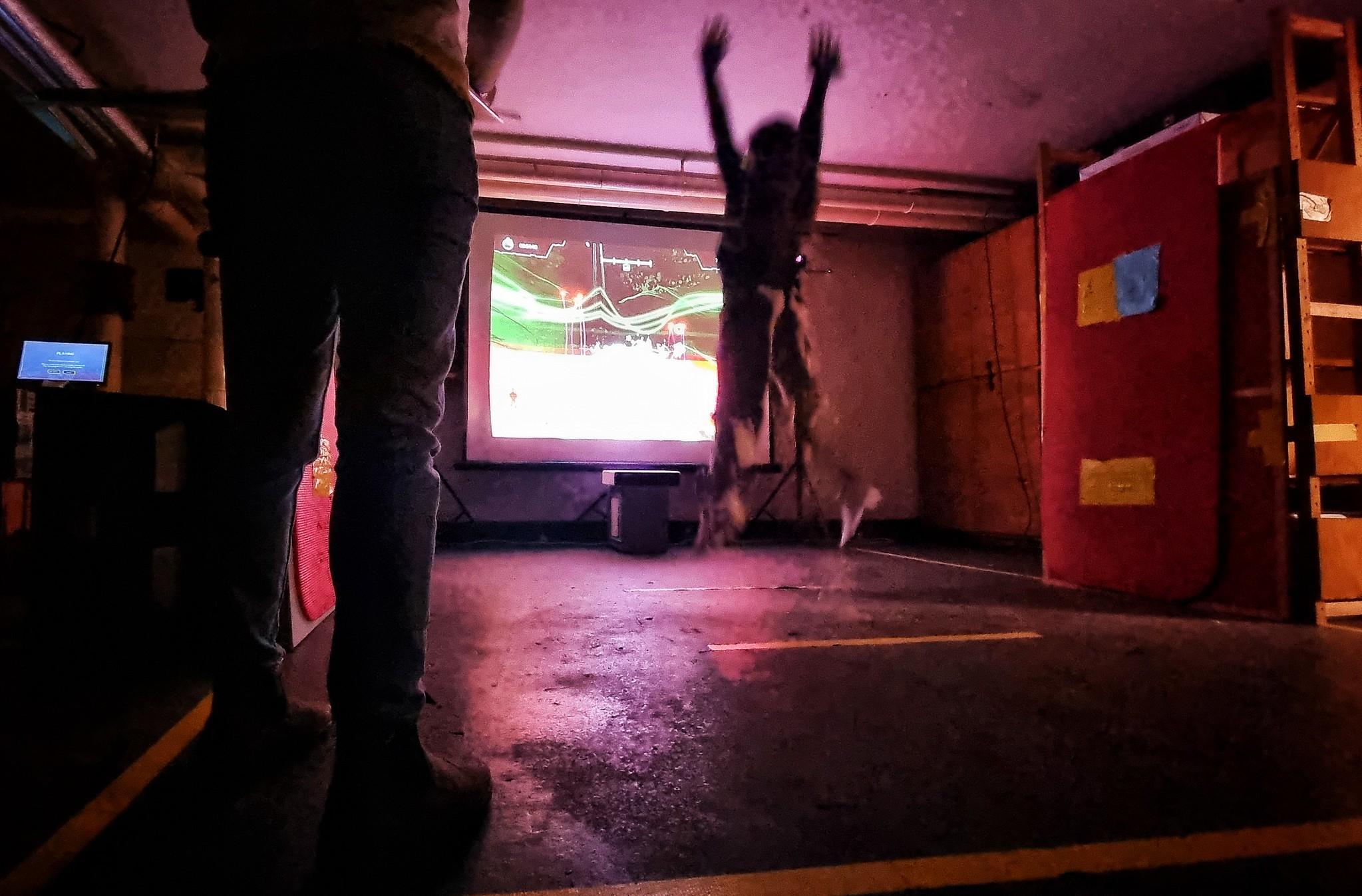 un enfant de dos est en train de sauter devant un jeu vidéo projeté sur un mur, tandis qu'un adulte l'observe