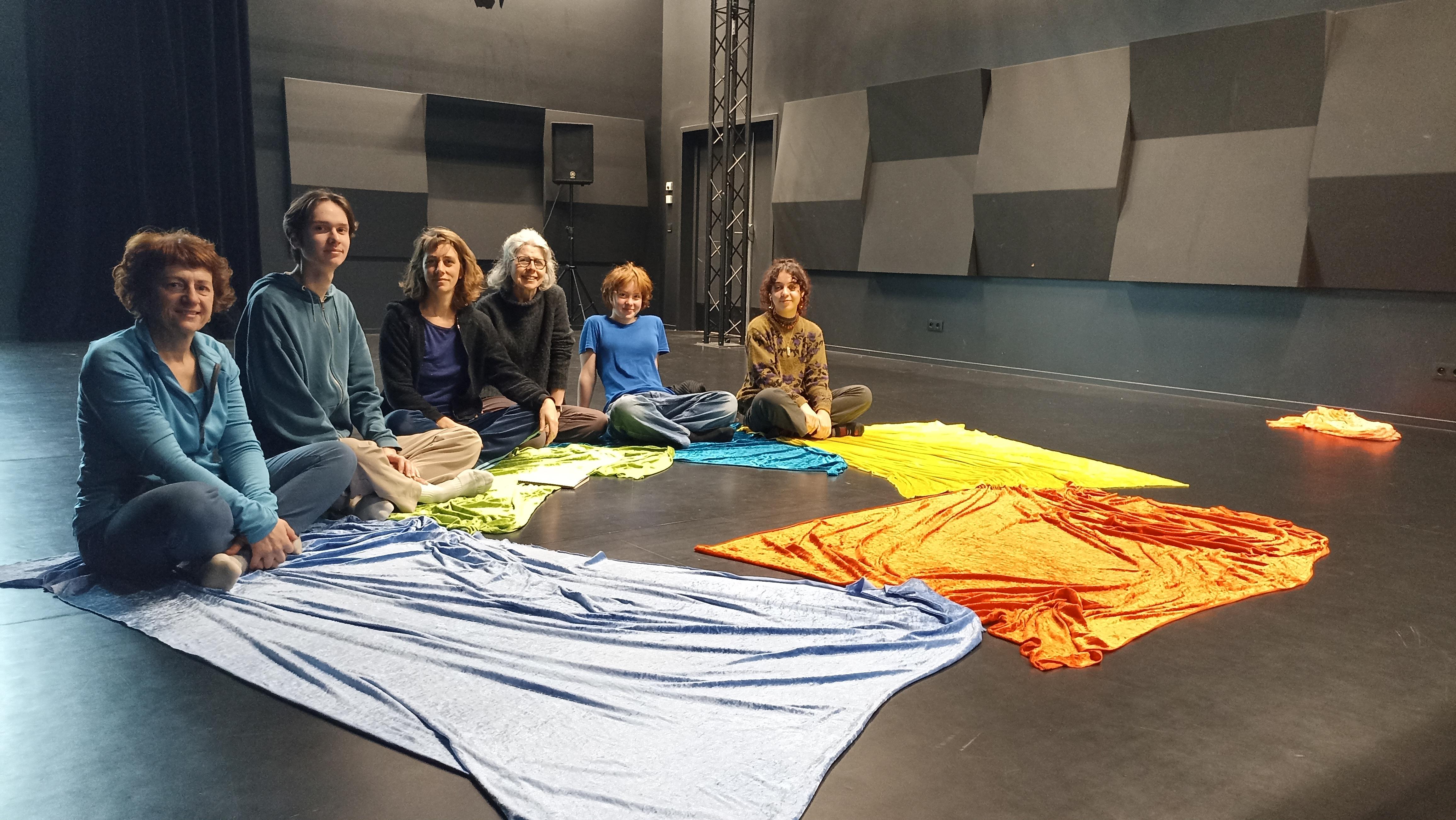 Des adultes et des enfants sont assis en cercle sur un sol de théâtre, entourées de grands morceaux de tissu coloré
