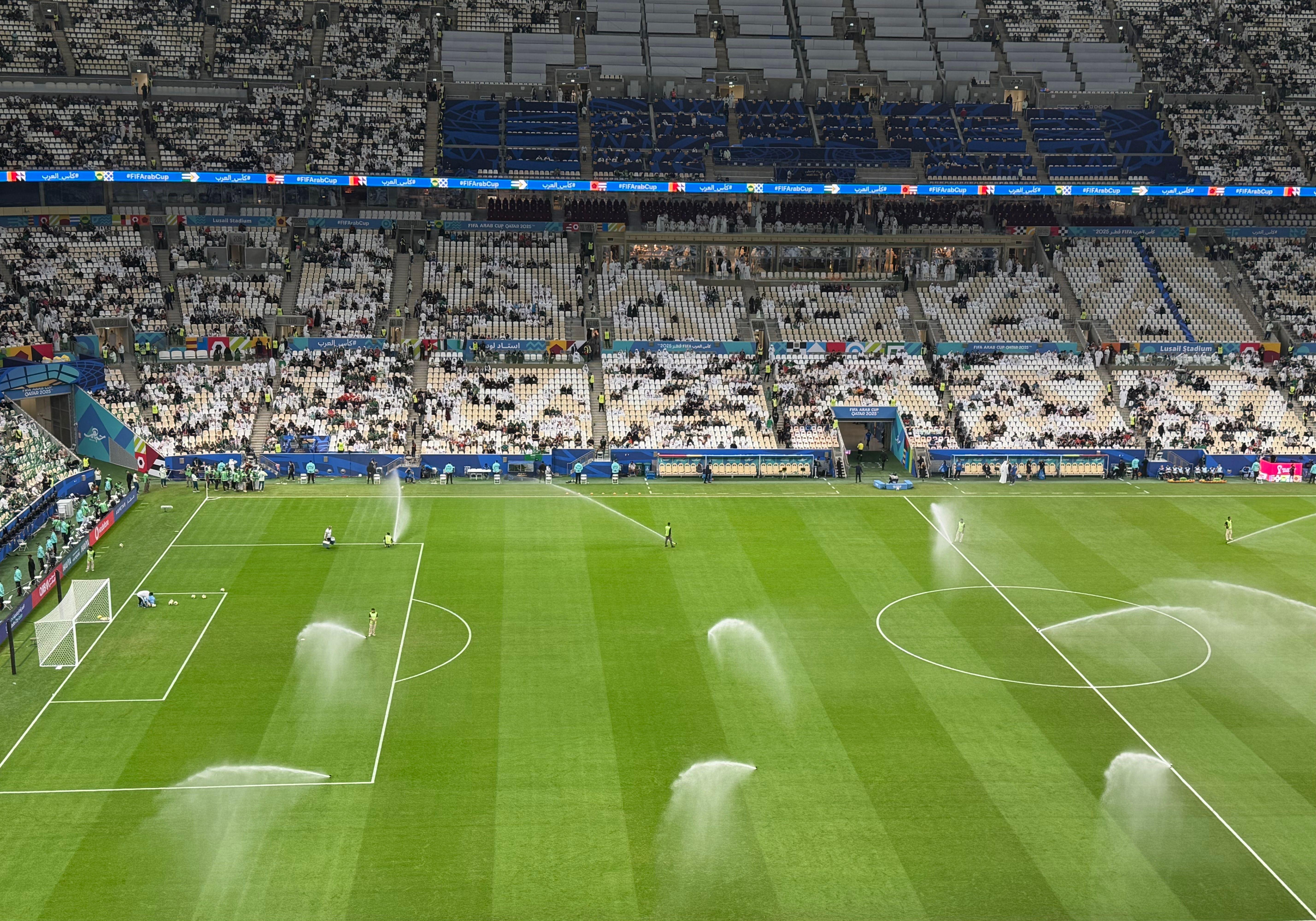 Arrosage du terrain d'un stade de football par des arroseurs automatiques sous un ciel nuageux en journée.