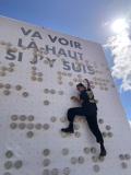 Une personne montre le mur braille, en haut du mur est écrit la phrase "Va voir là-haut si j'y suis"