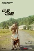Affiche du film "CRIP CAMP" Deux jeunes garçons sont au centre de l’image, sur une route d’un camp de vacances entouré de bâtiments blancs et de nature verdoyante. L’un est assis dans un fauteuil roulant, souriant, vêtu d’un t-shirt blanc et d’un pantalon noir. L’autre, debout derrière lui, torse nu et tenant une serviette sur l’épaule, pousse le fauteuil.