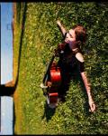 Photo de Dom La Nena dans l'herbe verte, allongée avec son violoncelle