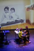 Photo issue du spectacle où on apperçoit deux comédiennes, brunes, asisses dos au public. Elles regardent la projection sur grand écran une photo de famille, sur laquelle deux petites filles sourient.