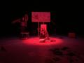 Photo prise lors d'une résidence de création, on y voit deux comédiennes sur scène, la llumière est sombre rouge. Un des comédiennes est au centre de l'image, elle lit un texte. L'autre comédienne se trouve derrière au fond de la scène