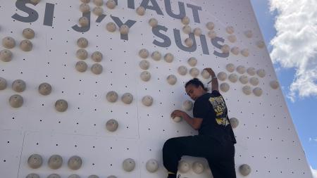 Une personne monte un mur braille, en haut du mur est écrit la phrase "Va voir là-haut si j'y suis"