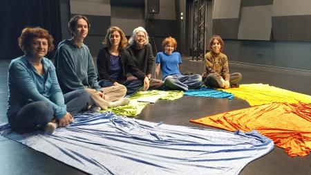 Des adultes et des enfants sont assis en cercle sur un sol de théâtre, entourées de grands morceaux de tissu coloré