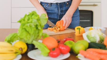 Mains d'une jeune femme en t-shirt blanc tranchant des légumes dans une cuisine moderne, se concentrant sur une alimentation saine.