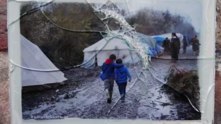 Photo d'un cadre en verre brisé contenant une photo de deux enfants qui marchent au milieu d'un camp de migrants, par Jeanne Menjoulet