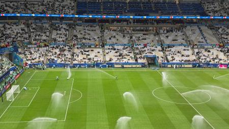 Arrosage du terrain d'un stade de football par des arroseurs automatiques sous un ciel nuageux en journée.