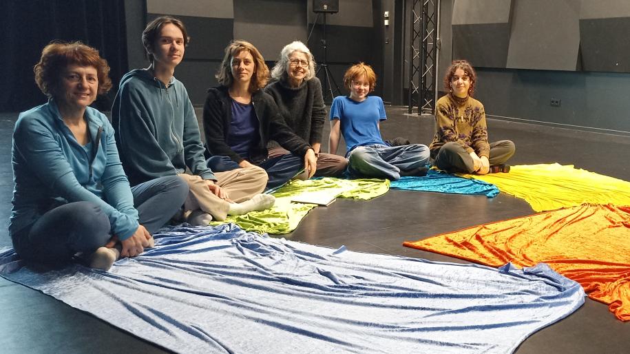 Des adultes et des enfants sont assis en cercle sur un sol de théâtre, entourées de grands morceaux de tissu coloré