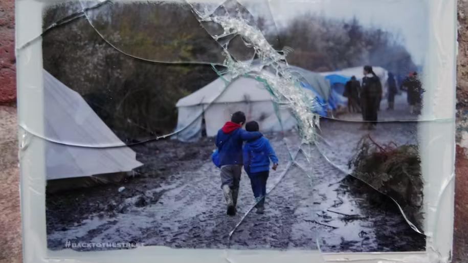 Photo d'un cadre en verre brisé contenant une photo de deux enfants qui marchent au milieu d'un camp de migrants, par Jeanne Menjoulet