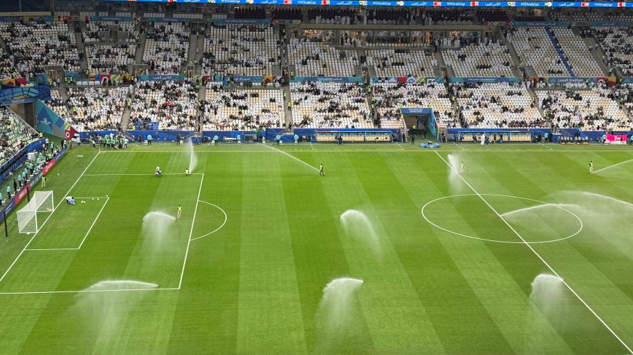 Arrosage du terrain d'un stade de football par des arroseurs automatiques sous un ciel nuageux en journée.