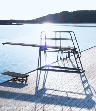 Photographie d'un plongeoire au bord d'une piscine avec en fond un ciel bleu et le reflet du soleil sur l'eau.