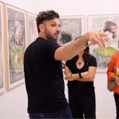 Photo portrait de Romuald Jandolo portant un t shirt noir des lunettes dans la main gauche devant une exposition