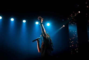 Une personne au cheveux long chante, un micro à la main et lève l'autre main, derrière on voit des lumière de scène bleu