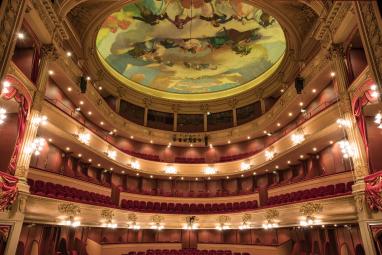 Intérieur d’un opéra à l’architecture classique, vu depuis la scène : plusieurs balcons ornés de dorures et de velours rouge entourent la salle, illuminés par de nombreux luminaires. Au plafond, une vaste fresque circulaire peinte représente des figures et des nuages dans des tons pastel.