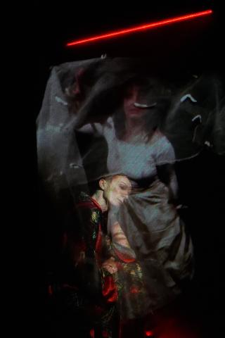 Visuel de "Rouge, amère fantasmagorie", on y voit deux figures humaines, créant un effet de flou et fantomatique. La personne à l'arrière-plan porte une robe blanche avec un visage partiellement visible, créant une impression de mouvement. Au premier plan, une autre figure est habillée de vêtements aux couleurs vives, principalement rouge et doré. La figure en arrière plan penche la tête sur le coté, donnant une impression de fragilité ou de réflexion.