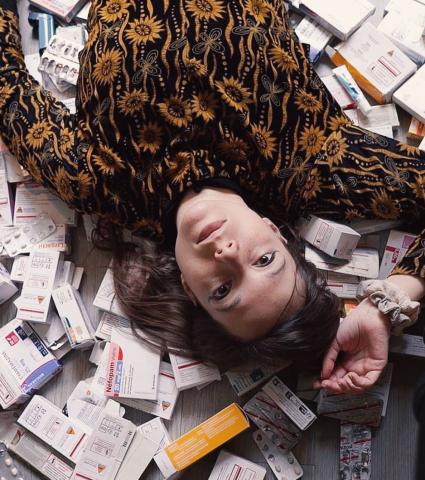 jeune femme allongée au sol, entourée de nombreuses boîtes et plaquettes de médicaments, regardant l'objectif