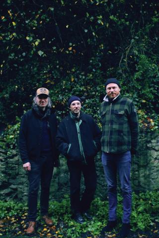 photo de trois hommes (membres du groupe), debout devant de la verdure (arbres et terre)