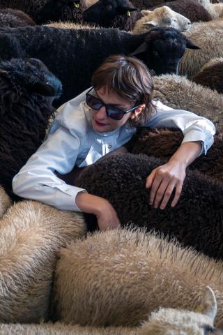 Visuel faire troupeau, photo de Marion Thomas, (au centre) portant des lunettes noires de soleil, entourée de d'un troupeau de moutons