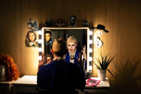 Une personne est assise de dos devant un miroir entouré d’ampoules allumées, dans une loge. Son reflet montre un visage transformé avec un maquillage drag très expressif, yeux soulignés de noir et lèvres accentuées. Elle porte une perruque beige sur la tête, en préparation d’une performance. Le miroir est décoré de découpages de portraits de stars (dont Elvis Presley et d'autres icônes hollywoodiennes).