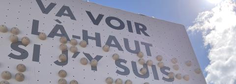 Une personne monte un mur braille, en haut du mur est écrit la phrase "Va voir là-haut si j'y suis"