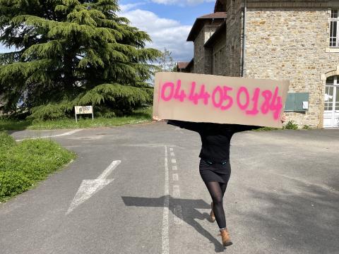 Céline Ahond en résidence artistique au Bois Perrin, portant un grand carton sur lequel est écrit en rose "0644050184", avec un bâtiment en pierre et des arbres en arrière-plan