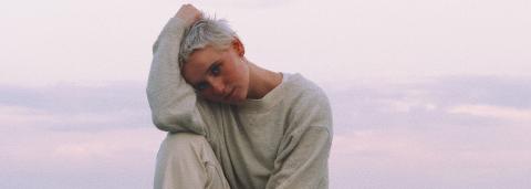 Kat White, e cheveux blond platine sur un rocher, au bord de la mer avec en arrière plan l'aurore, un ciel très claire, rosée