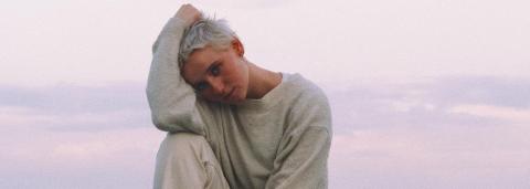 Kat White cheveux blond platine sur un rocher, au bord de la mer avec en arrière plan l'aurore, un ciel très claire, rosée