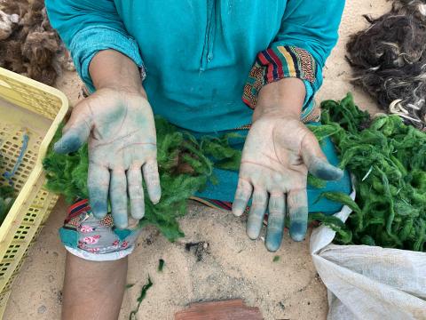 Sur la photo on voit une personne assise au sol, sur un terrain sableux, entourée de végétaux, les mains tachées, bleutées par la manipulation de ces végétaux. Elle porte une tenue colorée, bleue avec aux poignets des motifs mulicolores.